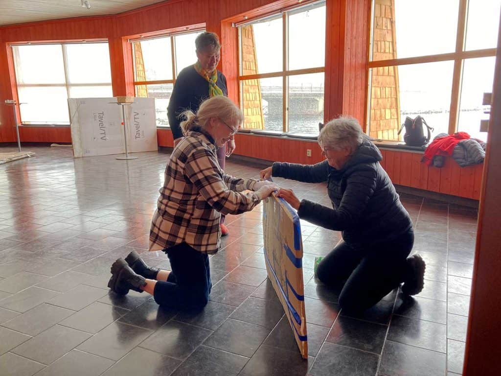 Två kvinnor står på knä på golvet och håller ett platt paket mellan sig för att tejpa ihop det. I bakgrunden står en tredje kvinna och tittar på.