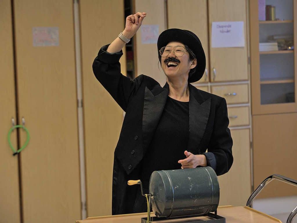 Glad kvinna med uppsträckt hand, utklädd med svart kostym, svart hatt och en mustasch. På bordet framför henne ett tombolahjul med vev.