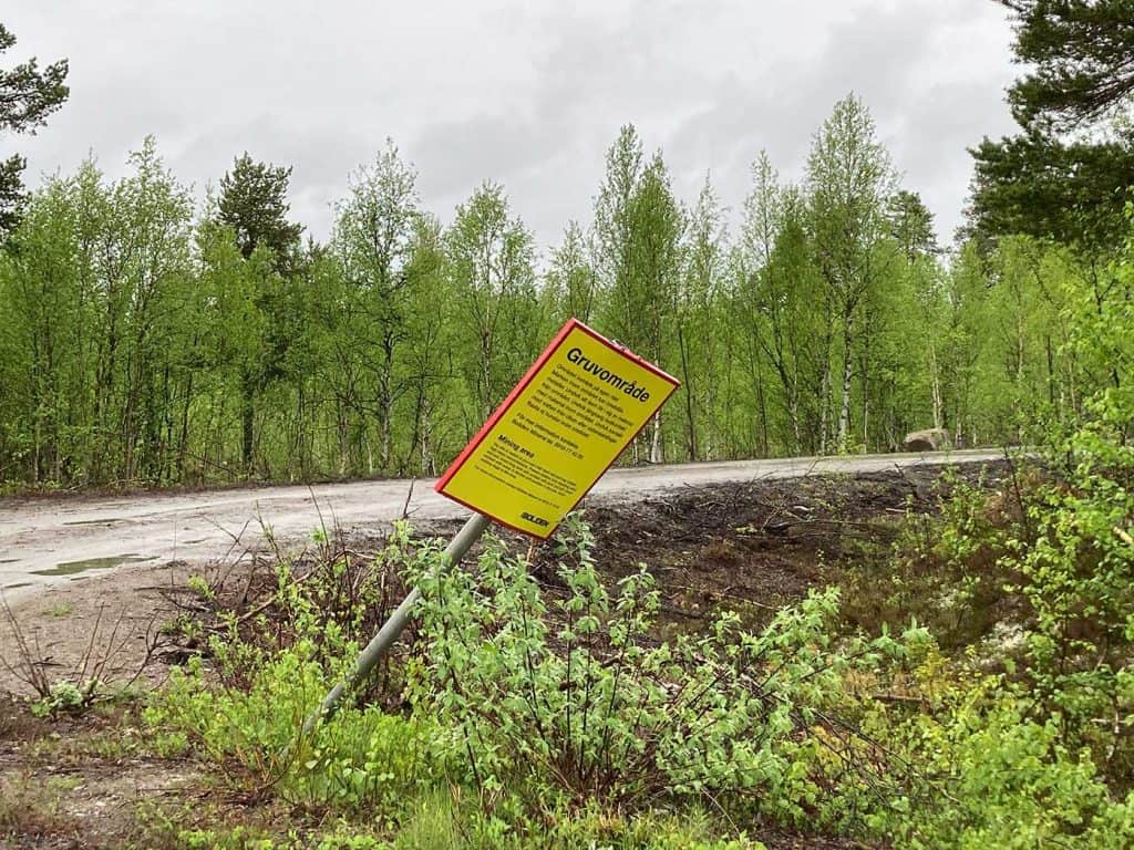 En lutande varningsskylt vid en grusväg i björkskog. På skylten sägs bland annat att gruvområdet beträds på egen risk och att man ska undvika att barn får i sig material från området.