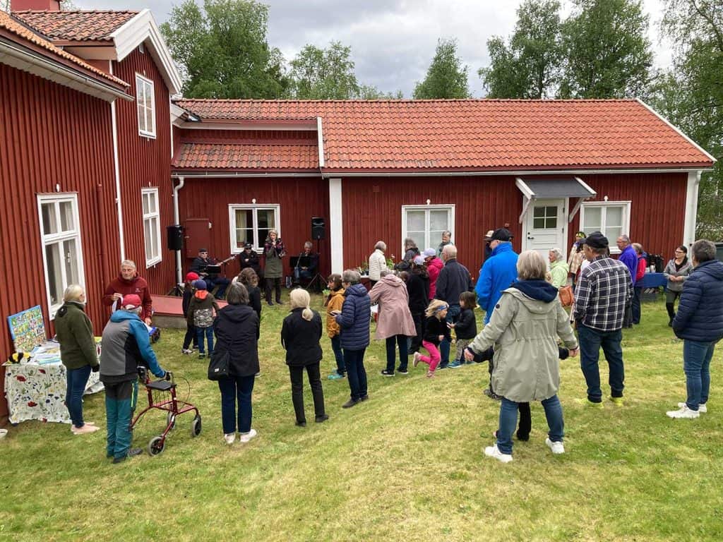 En översiktsbild med människor som står med ryggarna vända mot fotografen. De tittar mot en kvinna som håller tal. Bakom kvinnan syns tre musiker med sina instrument och ett gammalt hus, Doktorsgården.
