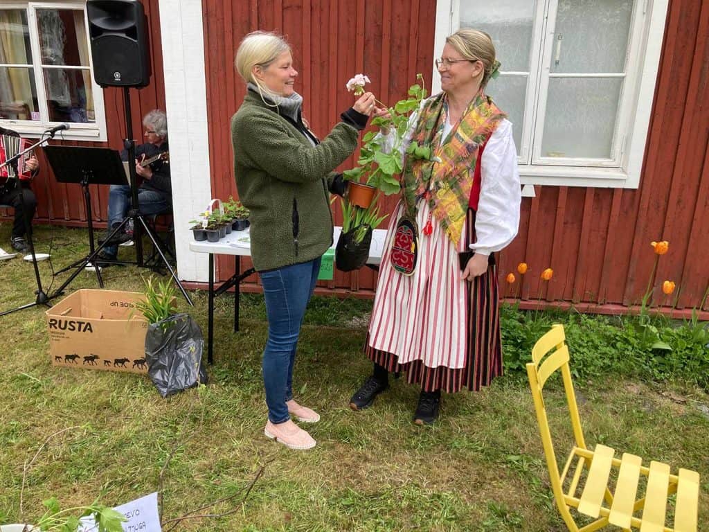 Två kvinnor står vända mot varandra. De håller en krukväxt, en pelargon, mellan sig, och ser glada ut. En av kvinnorna är klädd i folkdräkt.