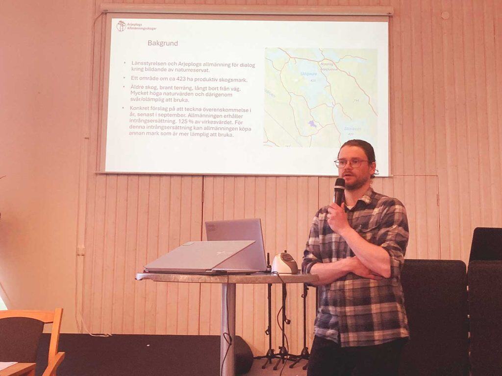 Karta och förklarande text om naturreservat på skärm i bakgrunden. En man med mikrofon i handen i förgrunden och intill honom ett bord med en bärbar dator.