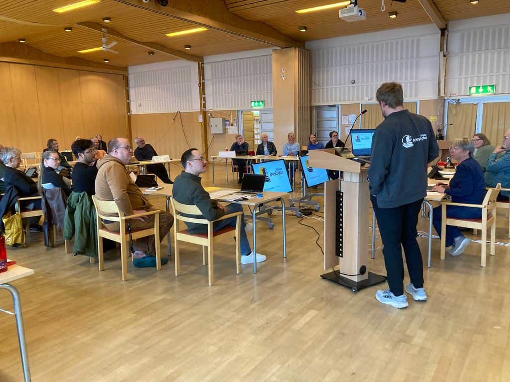 Bilden visar ett stort rum där mötesdeltagarna sitter vid bord och tittar på på en man som talar i mikrofon. Han står närmast kameran, med ryggen mot fotografen.
