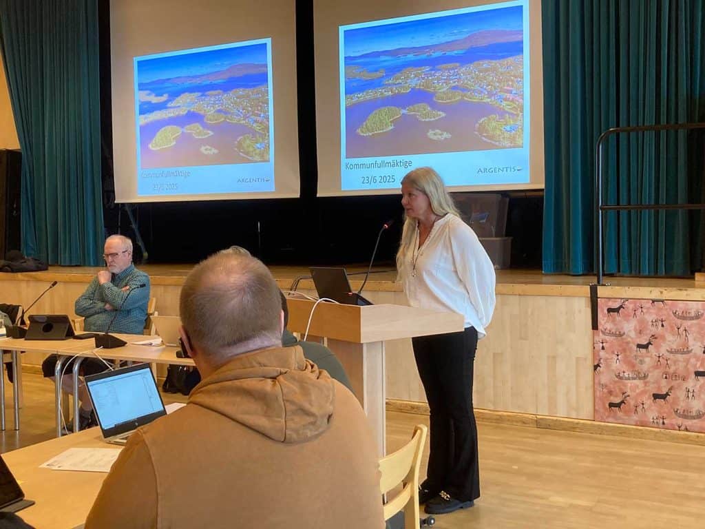 En kvinna vid en talarstol med mikrofon. Bakom henne två stora vita skärmar för bildpresentationer. På skärmarna visas i just detta ögonblick vybilder över Arjeplog.