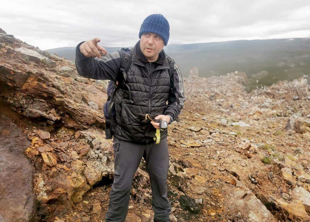 En man i täckjacka och toppluva, i ett stenigt område på kalfjället. Det är grått och mulet, men han ser glad och ivrig ut. 