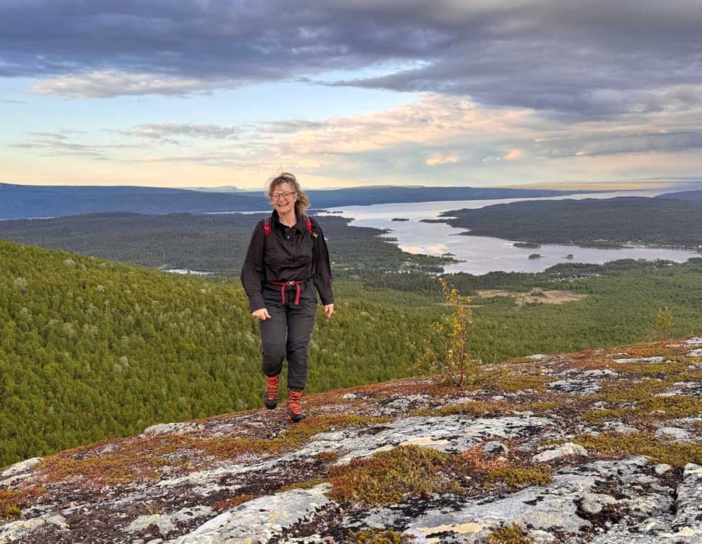 Ensam glad kvinna går mot fotografen uppe på ett berg. I bakgrunden syns skog och vatten.