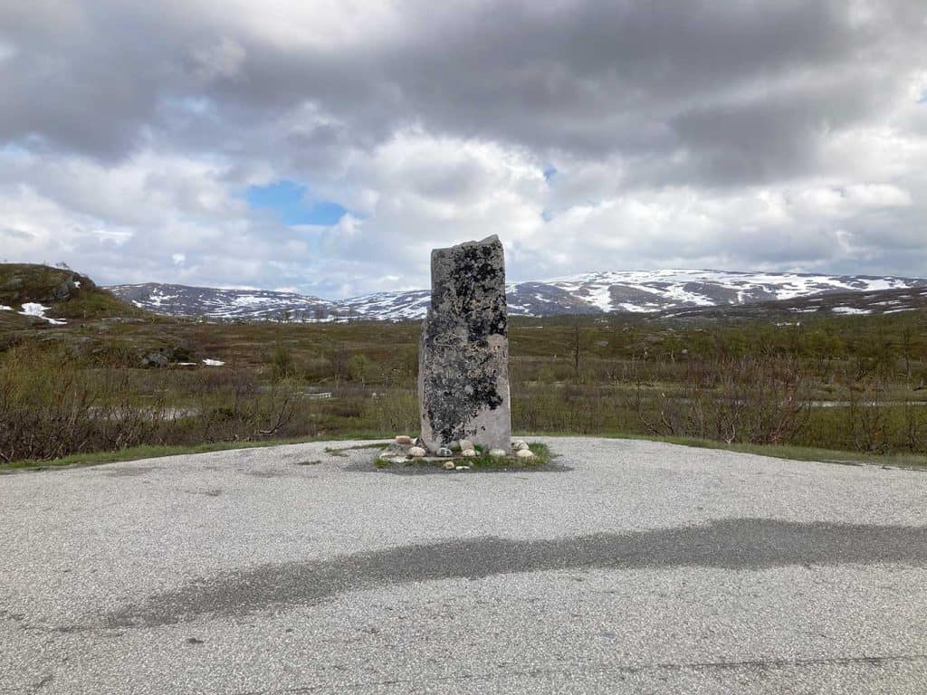 Bild på en stenstod på en asfalterad yta. I bakgrunden fjäll, delvis snötäckta. Blå himmel skymtar mellan regntunga moln.