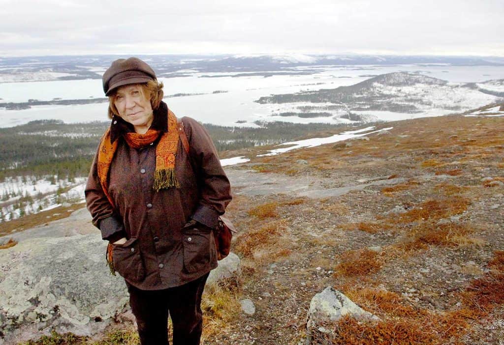 En kvinna med händerna i jackfickorna till vänster i bild. Hon står uppe på ett berg, och i bakgrunden syns vatten och skog. Det ser lite kyligt ut, med snö och is här och där.