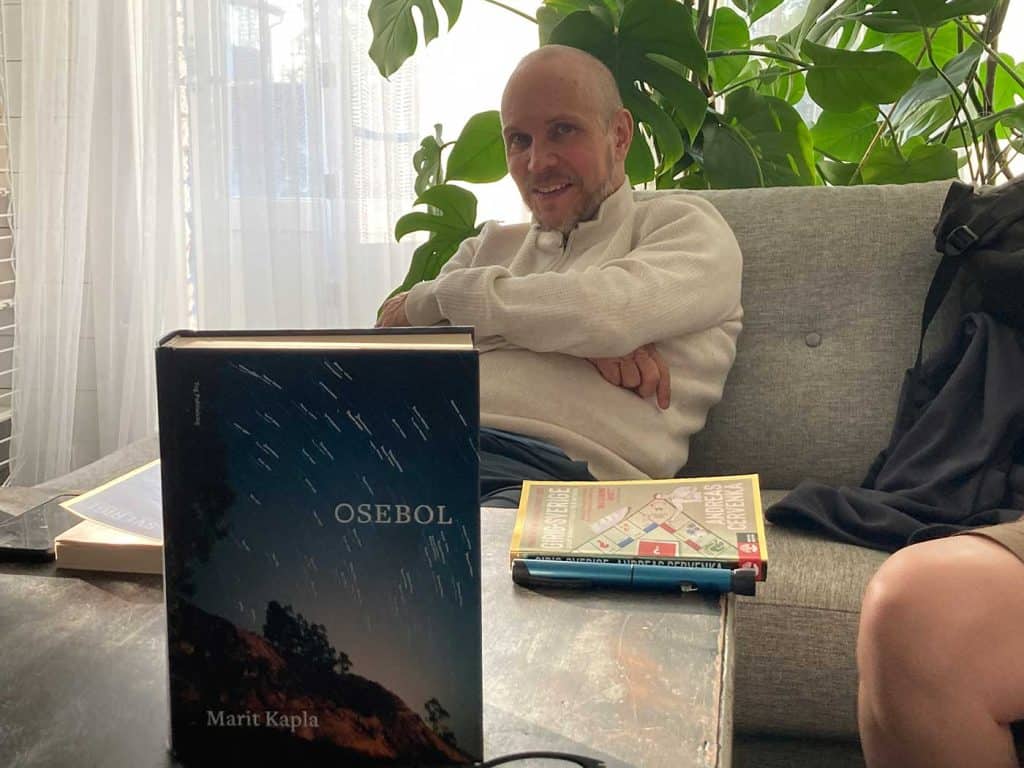 I förgrunden boken Osebol på ett soffbord. I bakgrunden sitter en man med korslagda armar och tittar leende på fotografen