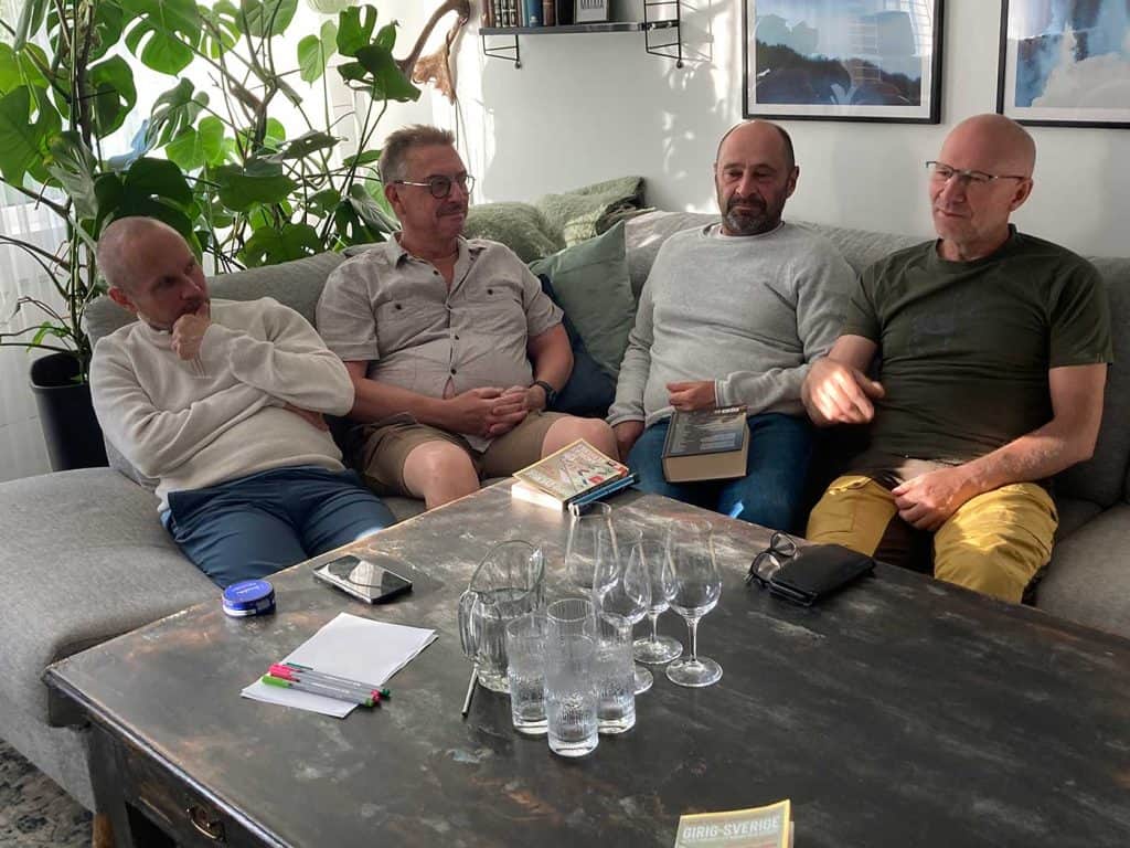 Fyra män sitter intill varandra i en soffa. På bordet framför dem finns en bok, en tillbringare, fyra vattenglas och fyra glas för whisky.