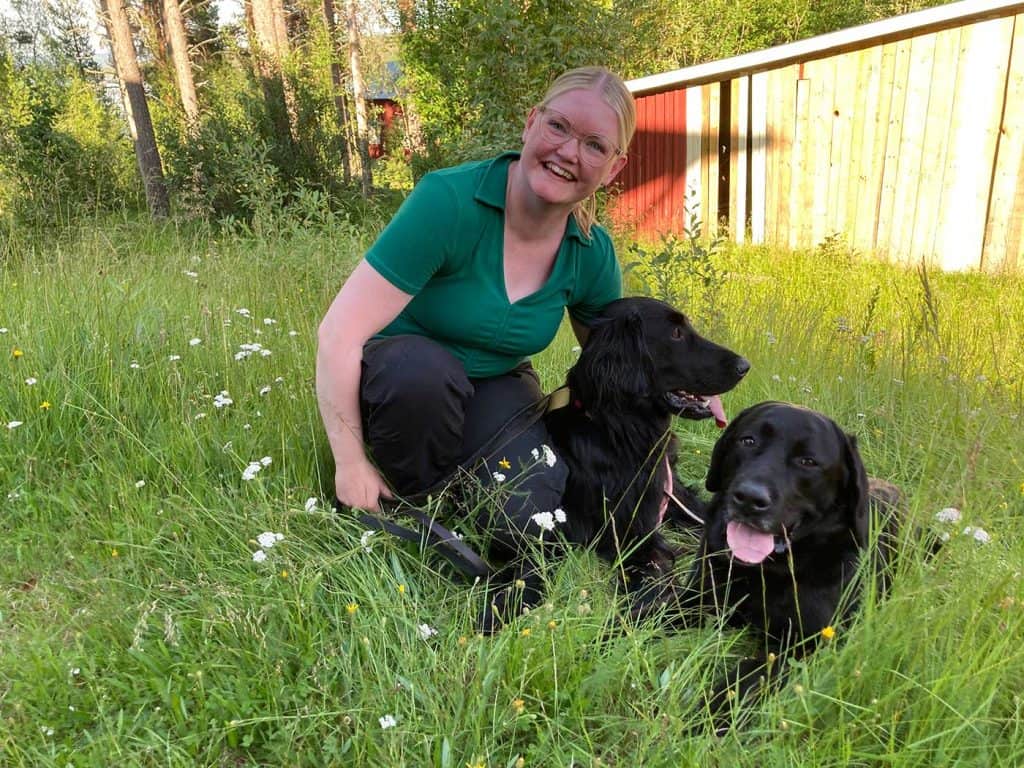Kvinna som sitter på huk i grönt gräs. Hon tittar på fotografen och skrattar. Bredvid henne ligger två svarta hundar. Deras tungor hänger ut ur öppna munnar.