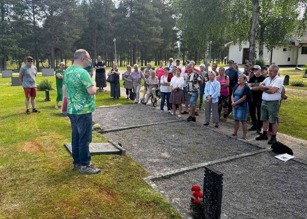 En grupp människor vid en grav på kyrkogården. Vid gravstenen, med ryggen mot fotografen, står en man som berättar om de människor som begravts här.