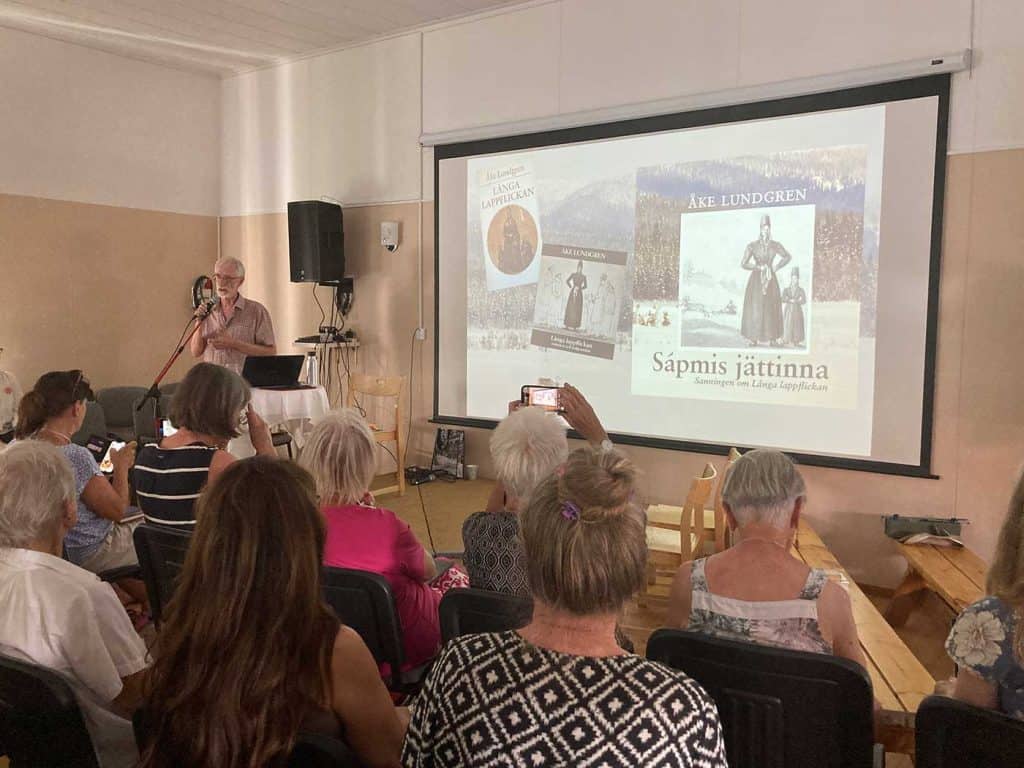 I förgrunden sittande publik, fotad bakifrån. Till vänster framför publiken står en man i kortärmad skjorta och talar i mikrofon. En stor del av bilden utgörs av en filmduk på väggen där man ser bilder av böcker om Långa lappflickan.