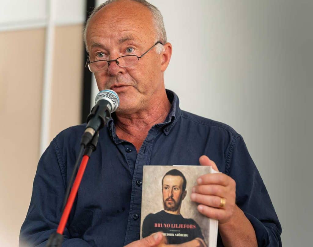 Man med glasögon står vid ett mikrofonstativ och pratar. Han håller upp en bok, endast den övre delen av boken syns i bild.