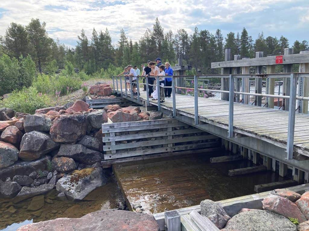 En grupp människor står vid ett broräcke och tittar ner mot vatten och stenar. Bron är också en del av en damm, och på bilden syns hur det kommer lite vatten genom dammluckorna.