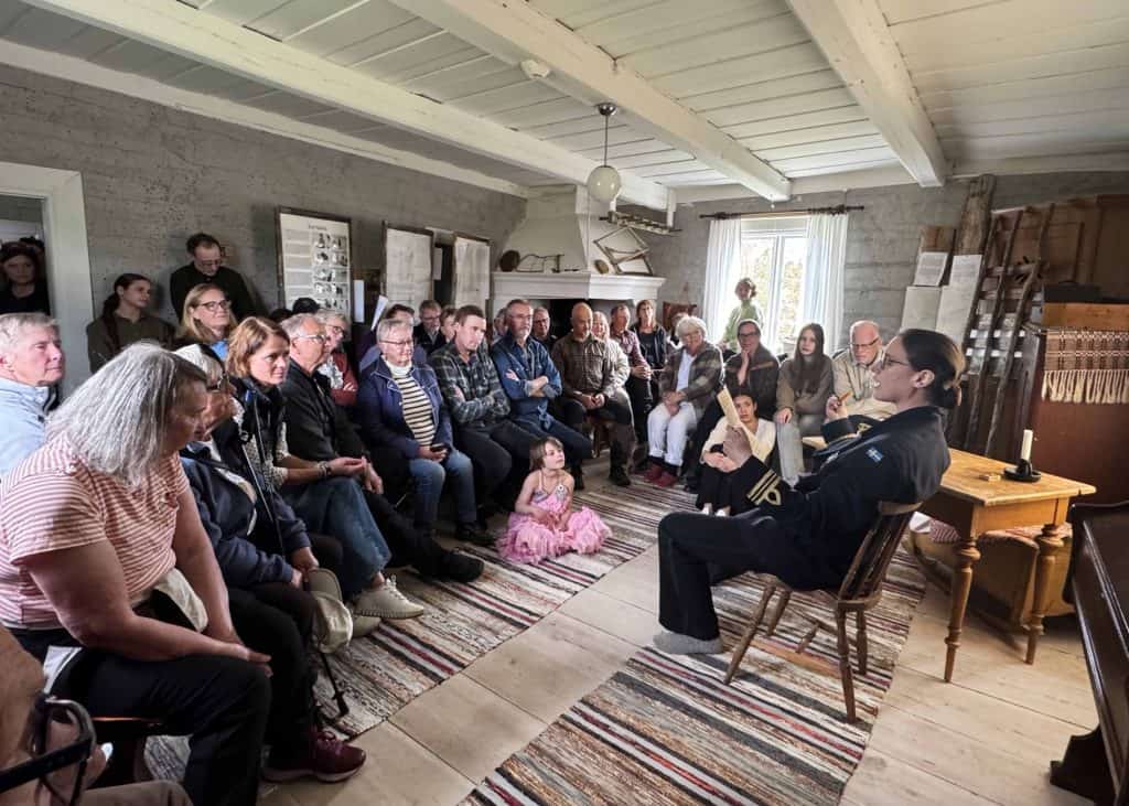 Människor sitter som publik på stolar, bänkar och golv i ett rum med timrade väggar, öppenspis och trasmattor på golvet. Till höger i bild sitter en kvinna på en gammal stol. Hon läser och gestikulerar.