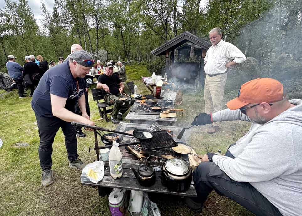 Två män lagar mat i stekpannor över eld. Vid elden står sotiga kaffepannor, och det är en del rök i bilden. I bakgrunden människor som tittar på.