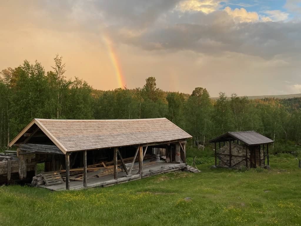 Ett gammalt sågverk under spåntak, i sommargrönska och med en regnbåge på himlen i bakgrunden. Till höger i bild den gamla masugnen, byggd av stenar, också den under tak. 