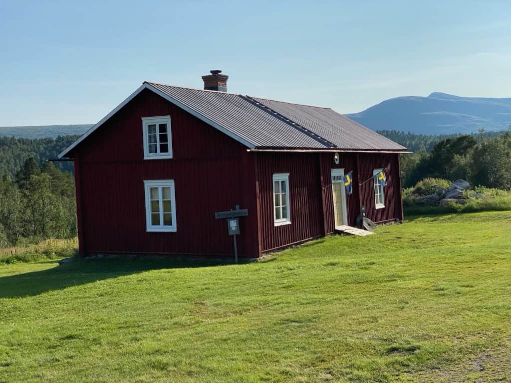 Ett rött timrat hus med vitt runt fönster och dörr. I bakgrunden ett fjäll och i förgrunden en grön gräsyta. 