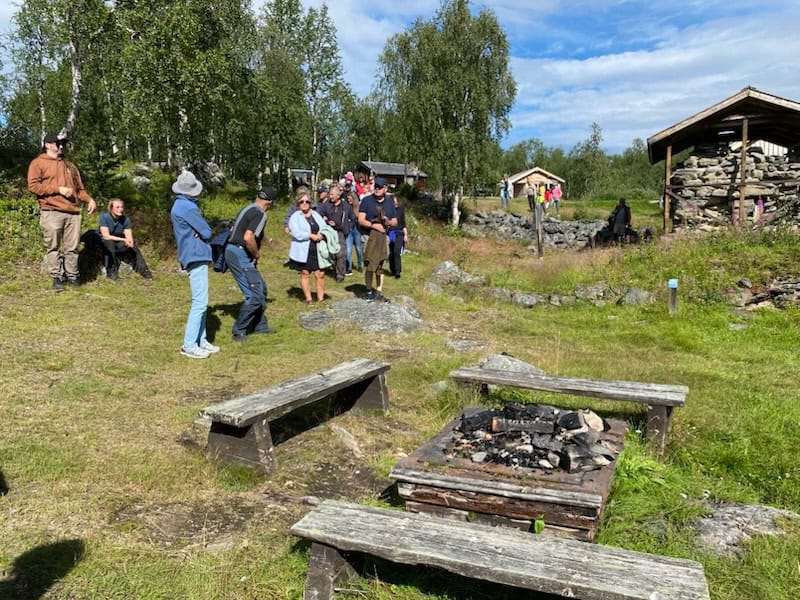 En sommarbild på en grupp människor som står och lyssnar när en person berättar om de rester av silverhyttan som syns, bland annat masugnen som byggts av stenar. I förgrunden en eldplats med sittbänkar runt om. 
