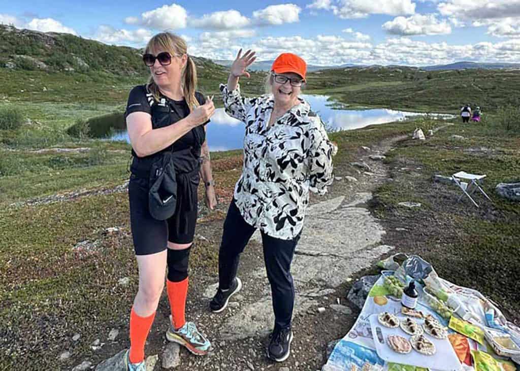 Två glada kvinnor i fjällmiljö. Bakom dem en liten tjärn, i övrigt kuperad fjällterräng med grön vegetation, samt en blå himmel med vita moln. En av kvinnorna är klädd för löpning, med kortbyxor, löparskor och ett knäskydd. Den andra kvinnan har långbyxor, en skjorta och keps.
