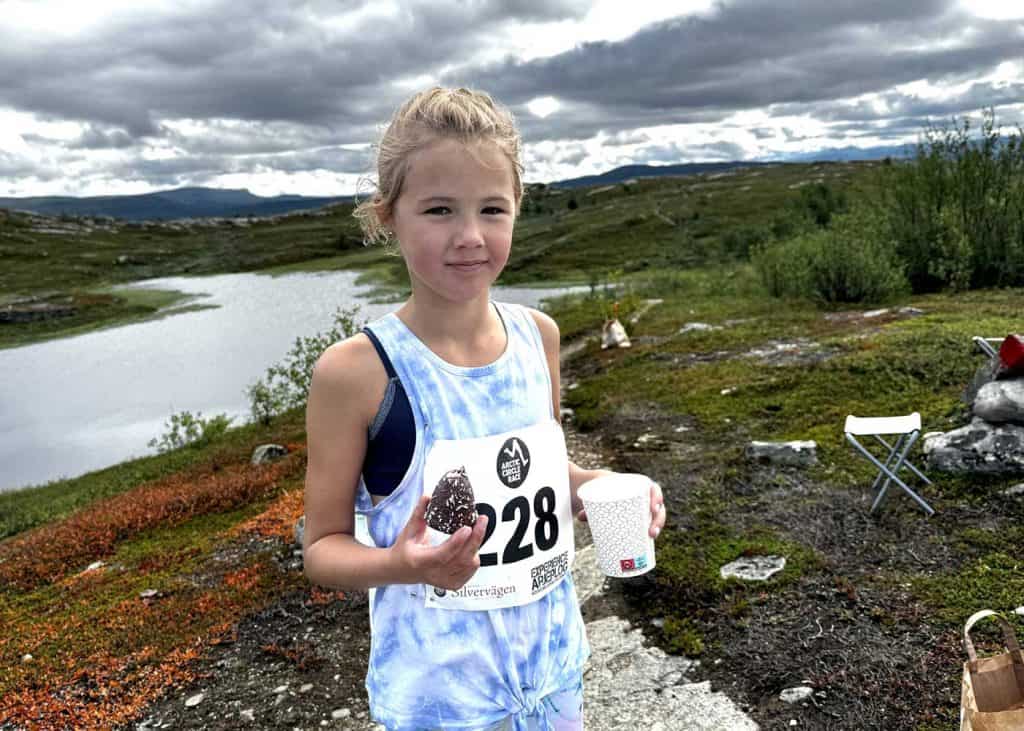Ung tjej i fjällmiljö, iförd träningskläder och en nummerlapp med siffrorna 228. I ena handen håller hon en mugg, i den andra handen en kokosboll. Hon har håret uppsatt och ser glad ut. 