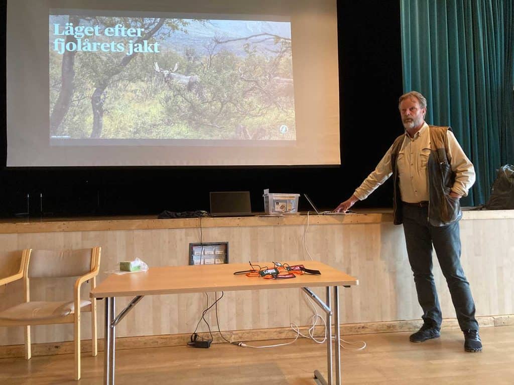 En man står framför en scen. Bakom honom en vit duk med projicerad bild av en älg i fjällmiljö med lågt björkris och rubriken ”Läget efter fjolårets jakt”.