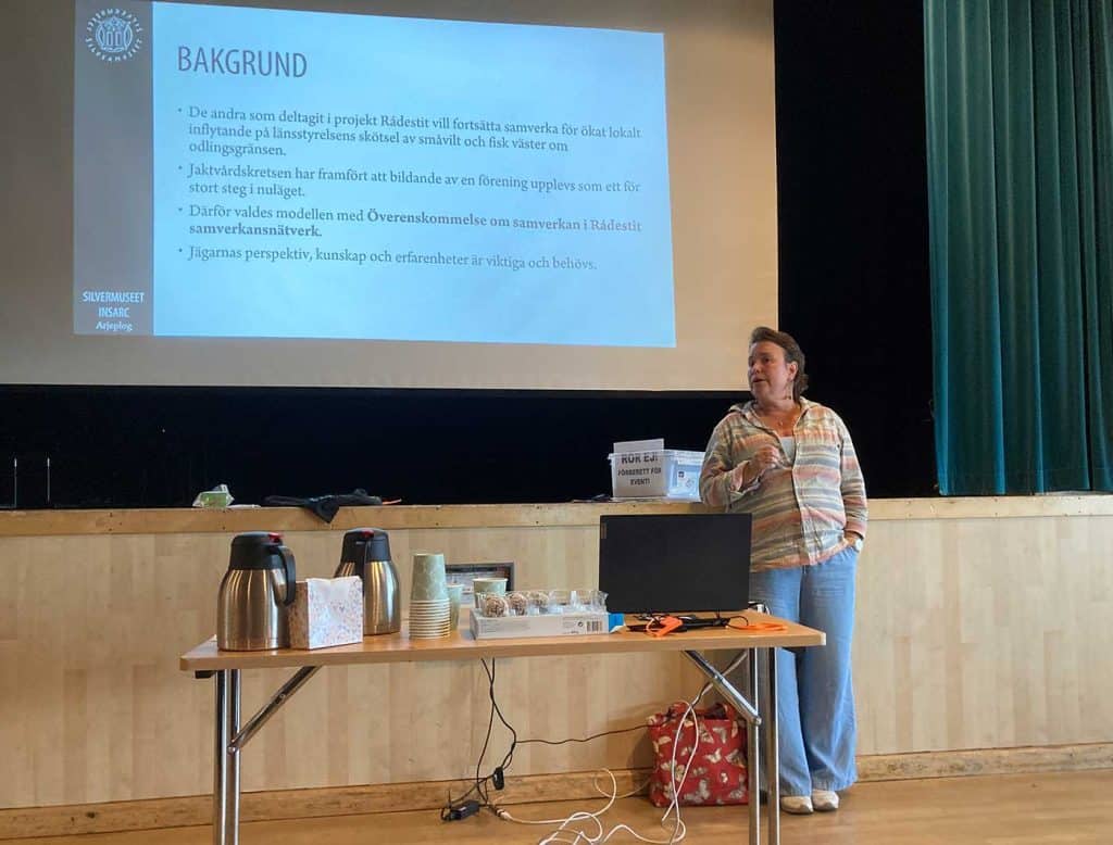 En kvinna står vid ett bord, framför en scen. Bakom henne en vit duk med en projicering av ett dokument med rubriken BAKGRUND och text som beskriver hur Jaktvårdsdkretsen har arbetat i projekt Rádestit.