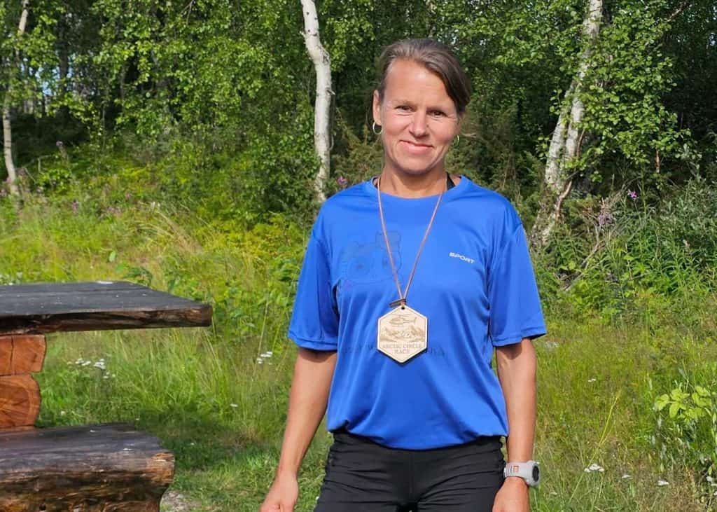 Glad kvinna med medalj runt halsen. Hon är klädd i en lös och ledig t-shirt. Omgivningen består av grön gräsmatta och björkar med gröna löv. 