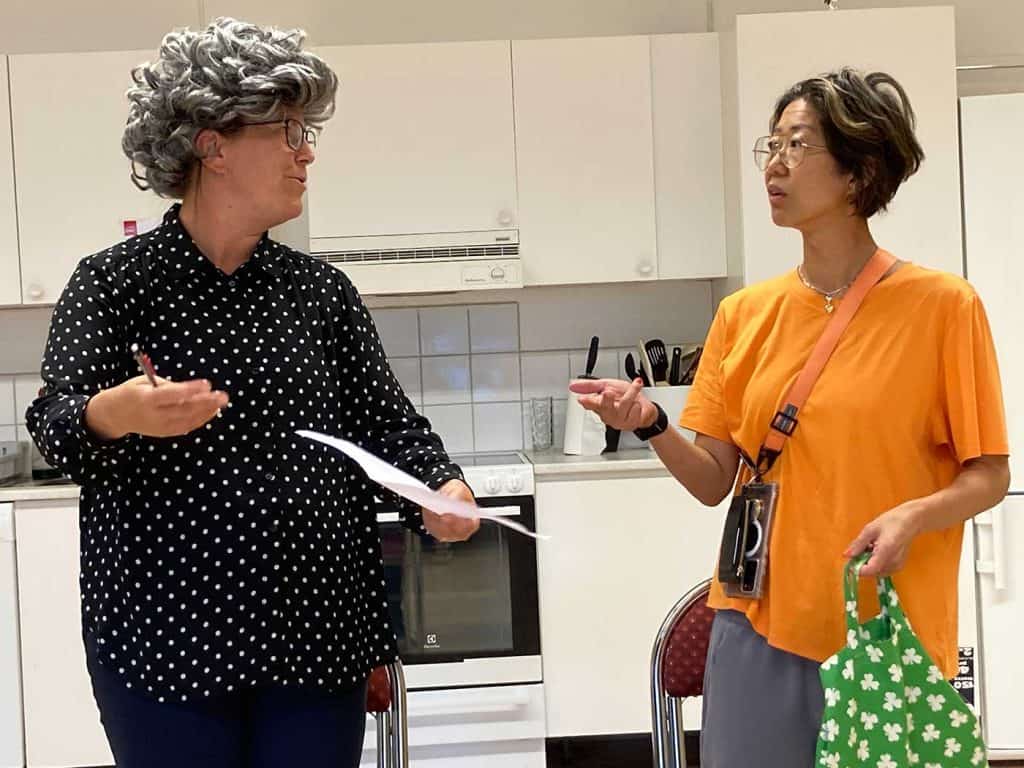 Kvinna i stor peruk till vänster pratar och gestikulerar. Till höger en kvinna med frågande ansiktsuttryck.