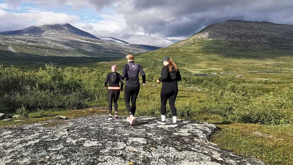 Fjällvy under molnig himmel. I bakgrunden fjälltoppar med lite snö på. Närmare fotografen är vegetationen låg men grön, till vänster i bild ett snår med fjällbjörk. Mitt i bilden syns tre kvinnliga löpare bakifrån, de springer över en flat stenhäll och deras hår, uppsatta i hästsvansar, fladdrar. Alla tre har svarta träningskläder och kepsar.
