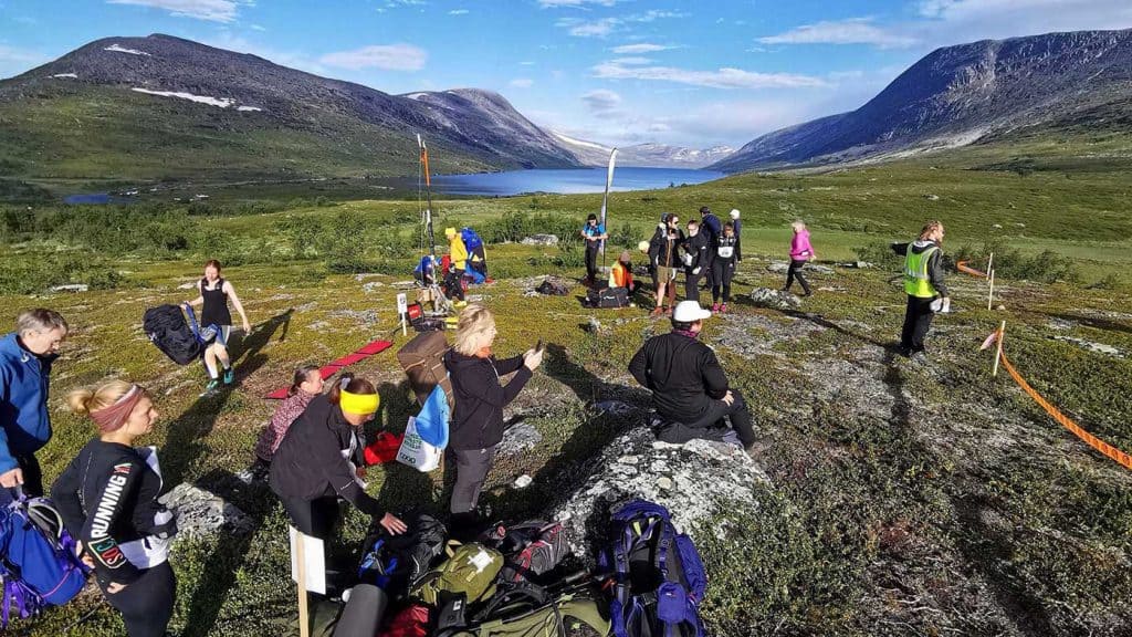 En vy med en sjö och fjäll med lite snö på topparna. I förgrunden, på kalfjäll med lite vegetation, människor i träningskläder som går, sitter och står i väntan på start. Det finns också en samlad hög med ryggsäckar i förgrunden.