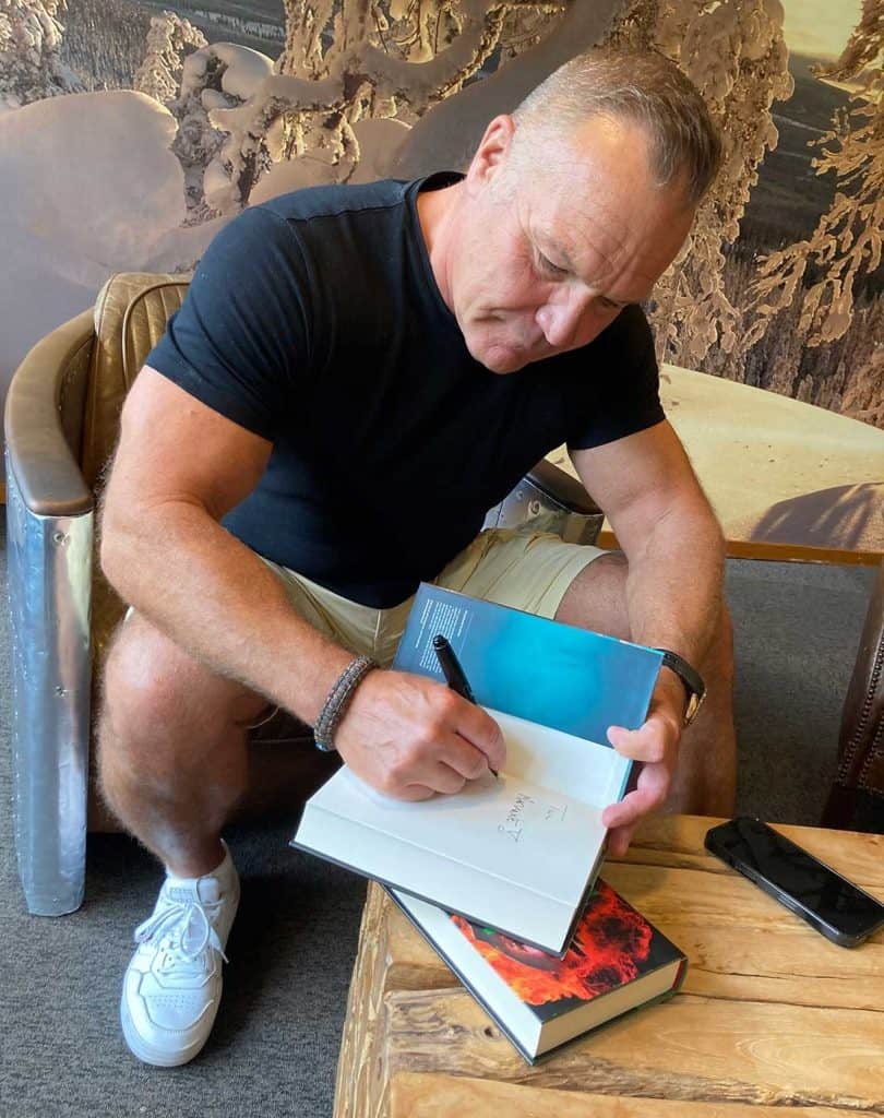 En man sitter i en fåtölj, lutar sig fram mot bordet och skriver en hälsning i en bok.