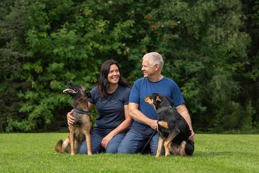 Bild på en kvinna och en man som sitter på en gräsmatta. De har varsin hund bredvid sig och ser glada ut.