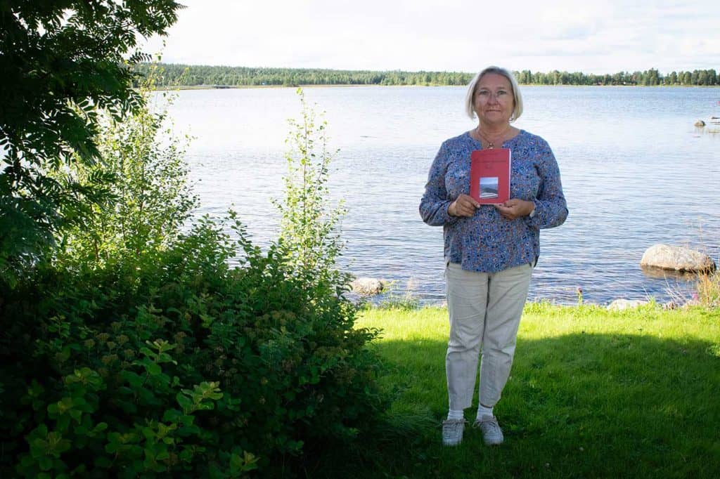 En kvinna i helfigur, håller upp en bok framför sig. Hon står utomhus, det är sommar och mycket grönska, vatten i bakgrunden