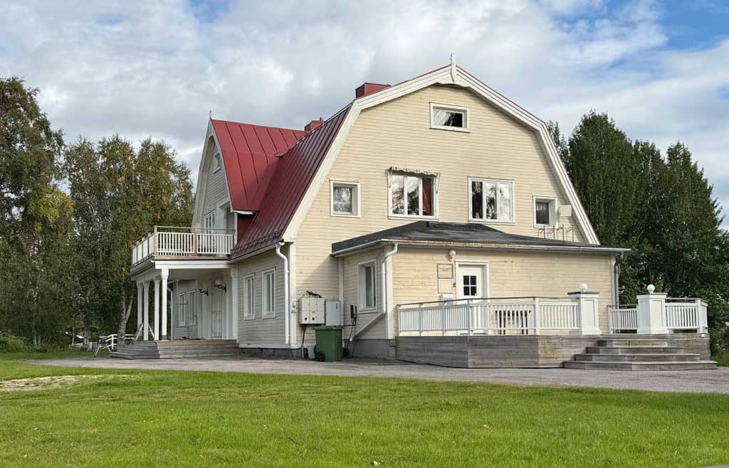 Ett stort hus, fotat från ena kortsidan. Där finns en lägre utbyggnad med en ytterdörr och bro. På långsidan, som också är husets framsida, ser man en bro där brotaket samtidigt är en balkong. 