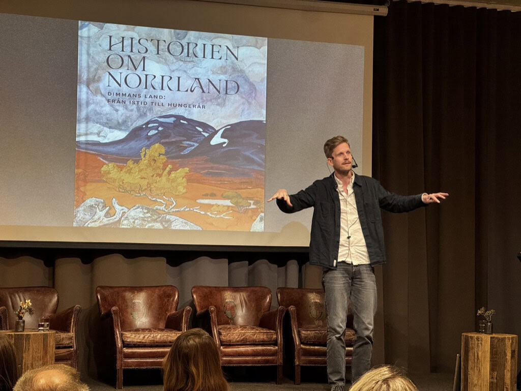 En man på en scen, han gestikulerar och håller ut armarna från kroppen. Bakom honom på väggen ett projicerat bokomslag. Bokens titel är Historien om Norrland, Dimmans land från istid till hungerår.