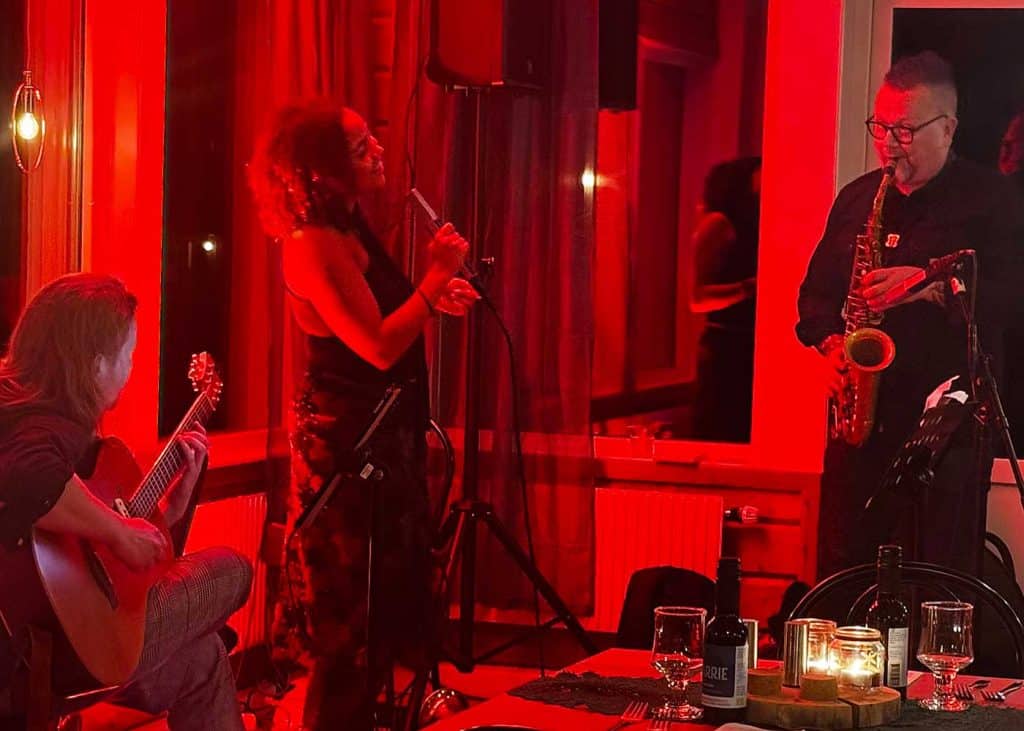 En kvinna står med mikrofon i hand. Hon ser glad ut och riktar uppmärksamheten mot en man som spelar saxofon. Bilden badar i rött ljus och man kan se ett bordshörn med glas och flaskor på.
