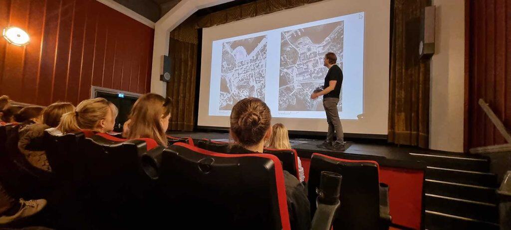 Bilden visar en stor filmduk med bilder av Arjeplogs tätort. Till höger i bild står en man och tittar mot filmduken. I förgrunden syns delar av publiken bakifrån.