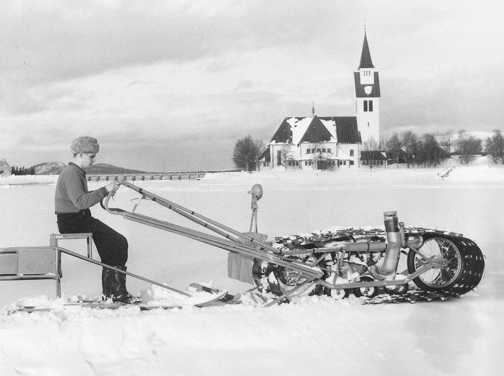 Gammal bild på en man som kör ett fordon på is. I bakgrunden Arjeplogs kyrka.