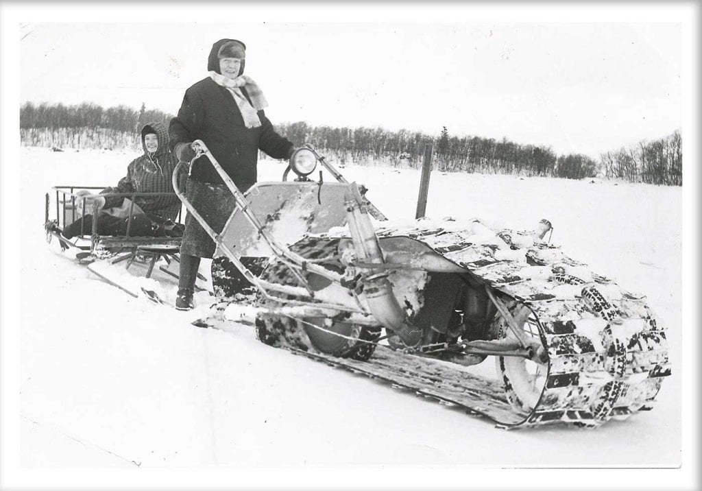 Gammal bild med en kvinna som kör ett snöfordon stående. Hoh ser glad ut. På en kälke bakom sitter en annan kvinna som också ser glad ut.