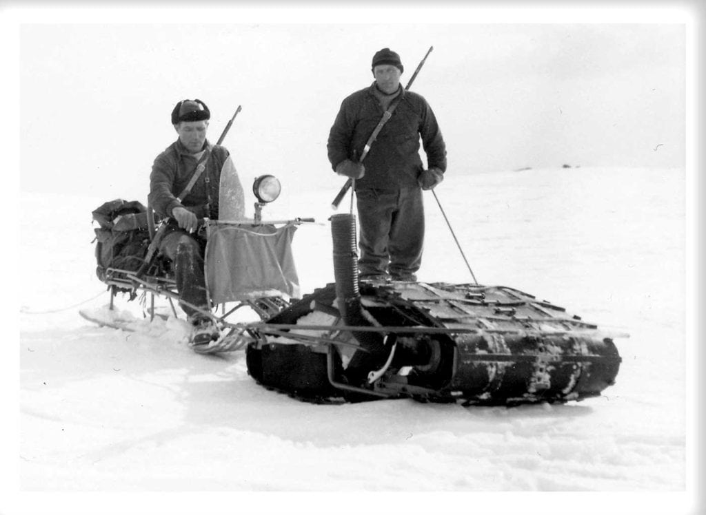 Gammal bild på två män, bägge har gevär med sig. Den ene sitter på ett fordon, den andra står bakom fordonet med skidstavar i händerna. 