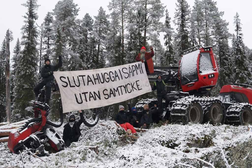 Bild på skogsmaskin och människor vid och på maskinen. De håller upp en skylt med texten ”Sluta hugga i Sápmi utan samtycke”