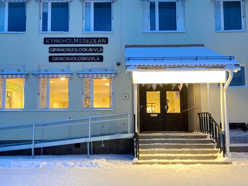 Vinterbild på en entre med brotrappa, dubbeldörrar med glasrutor och ett brotak. På vänster sida av bron en rullstolsramp, och skyltning Kyrkholmsskolan.