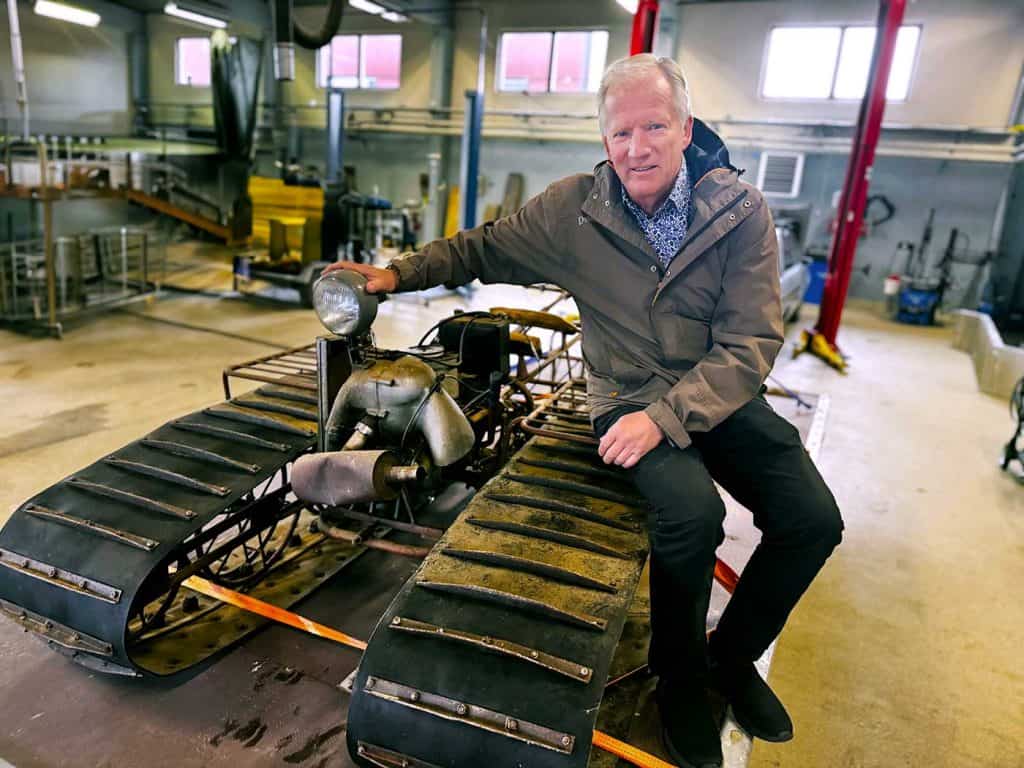 En man sitter på ett snöfordon i ett garage. Det är ett gammalt snöfordon, föregångare till snöskotern.