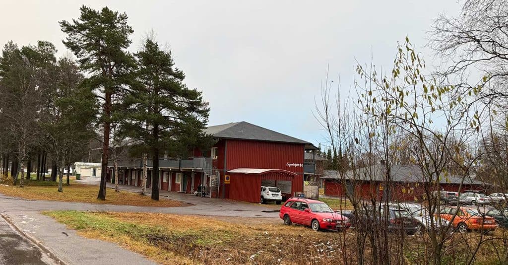 Ett hyreshus i två våningar. Utanför huset står ett par bilar parkerade. 