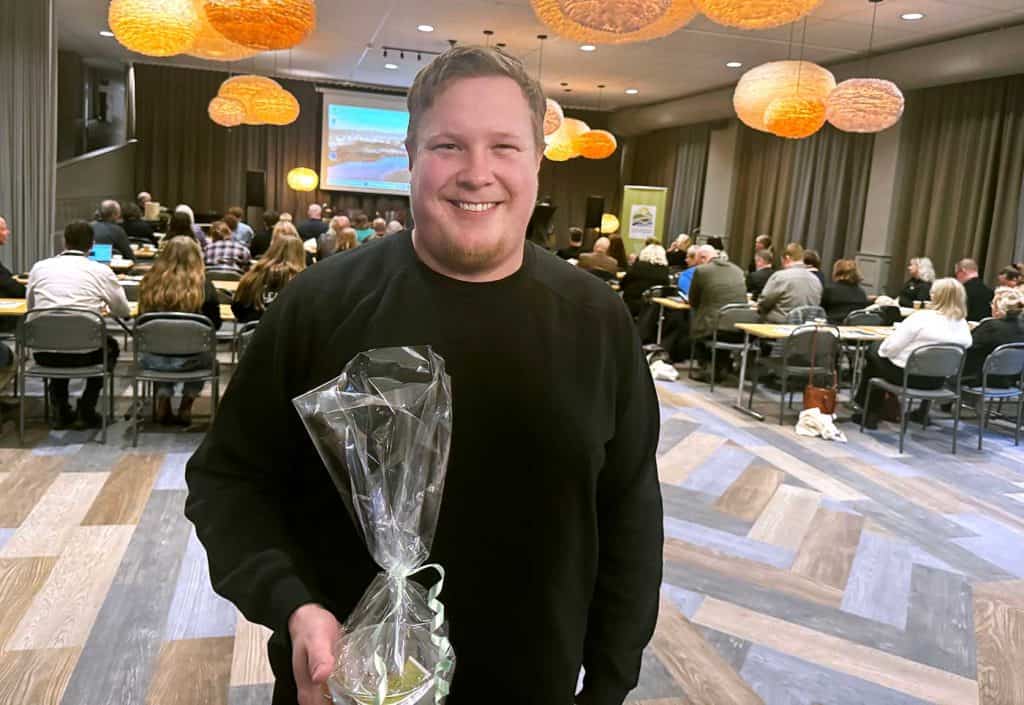 En glad man i en konferenslokal. Bakom honom sitter folk vid bord, med ryggarna mot fotografen. Deras uppmärksamhet riktas mot föreläsaren längst fram i salen.