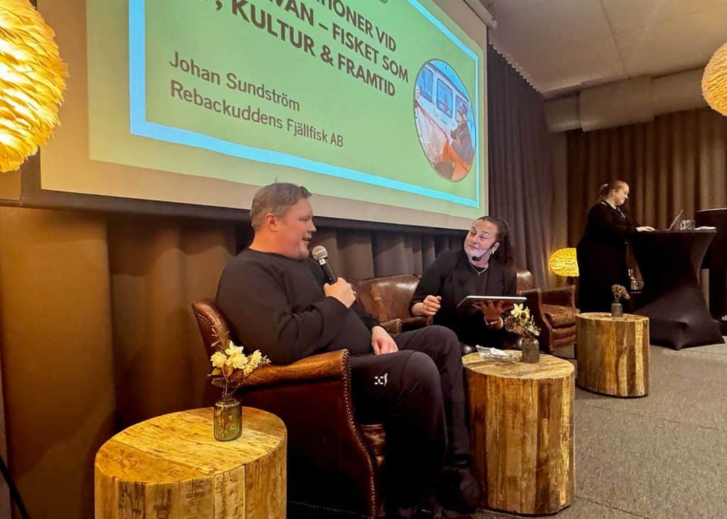 En man och en kvinna sitter i en soffa. Mannen håller i en mikrofon och kvinnan har headset på sig. På väggen bakom dem en skylt där man kan se en del av texten: Fisket som kultur och framtid, samt namnet Johan Sundström, Rebackuddens Fjällfisk AB.