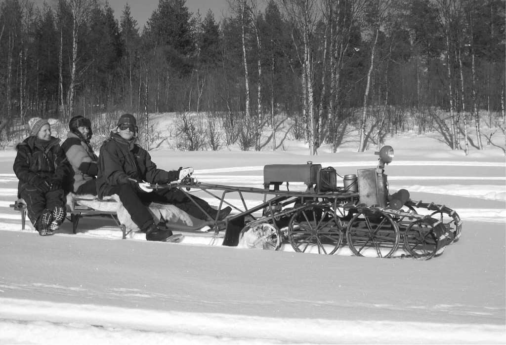 En vinterbild i svartvitt, vilket ger ett lite gammalt intryck. Tre personer sitter på ett snöfordon. De ser glada ut. 
