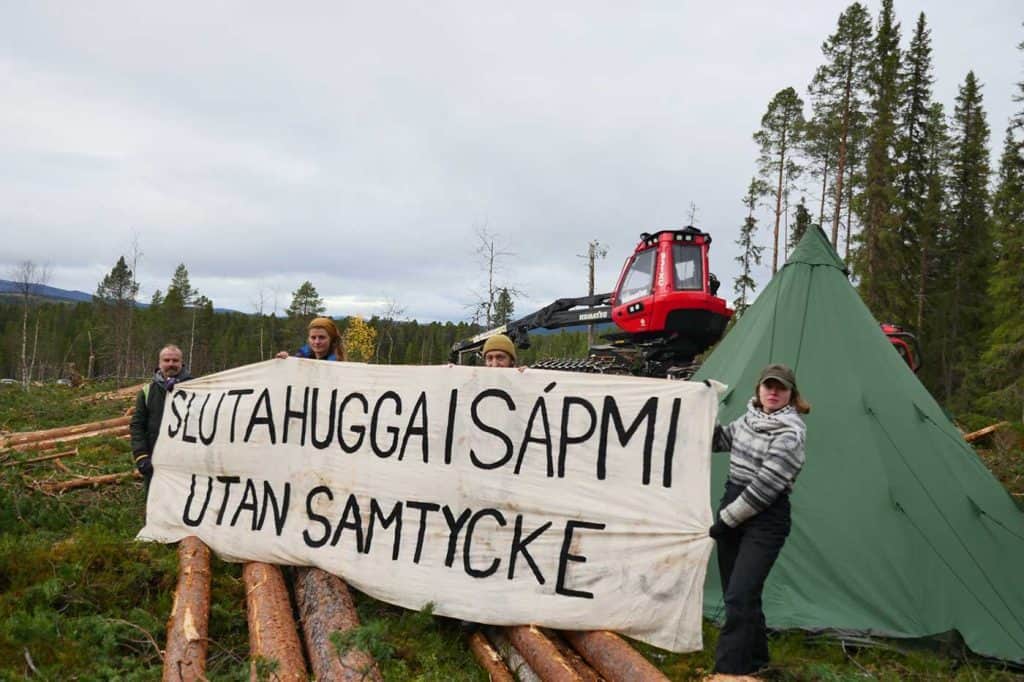 Bild från ett avverkningsområde i skogen. Fyra personer står vid en skogstraktor och fällda träd. De håller upp en skylt med texten